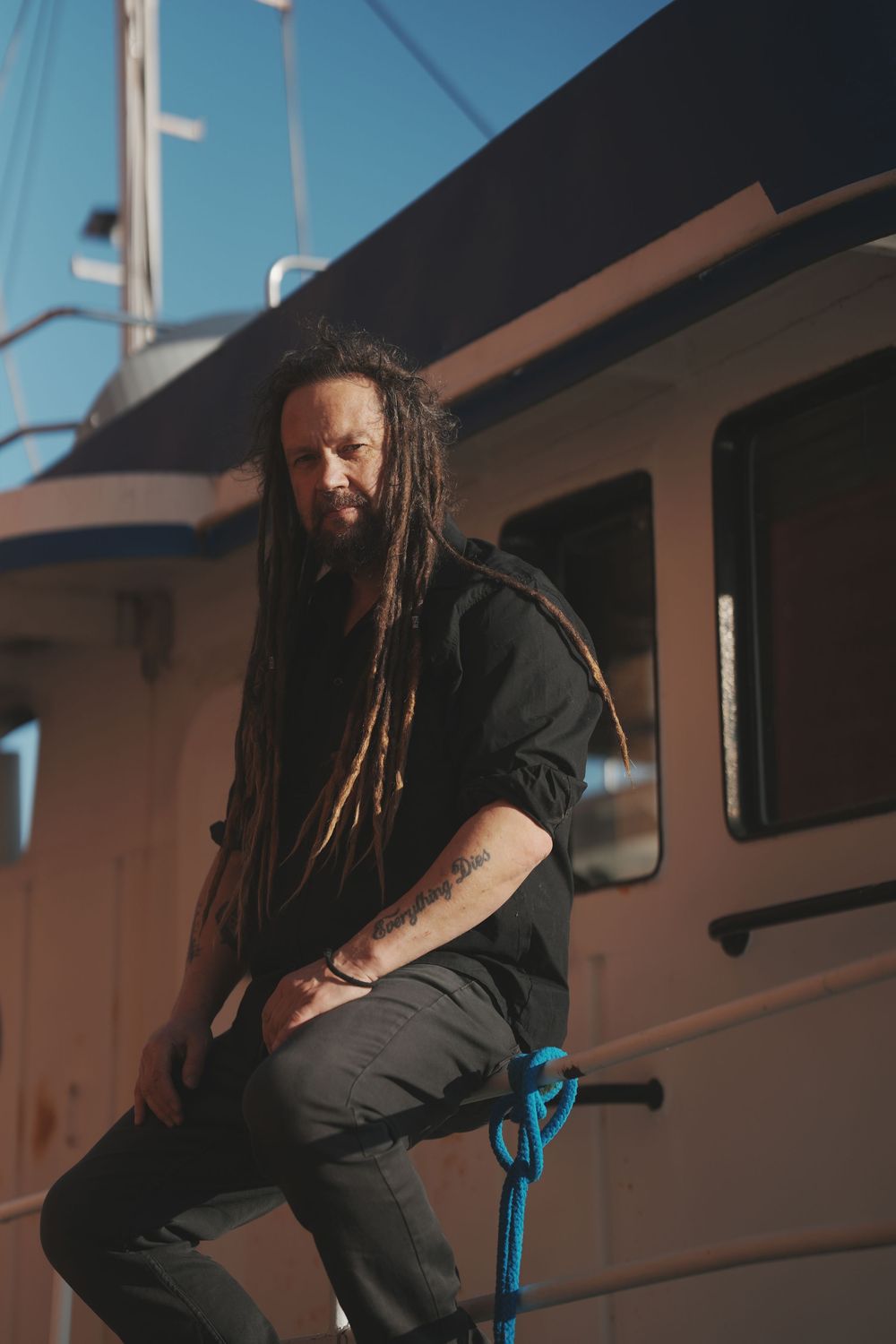 Person with long dreadlocks wearing dark clothing sits casually on a boat against a bright sky background.