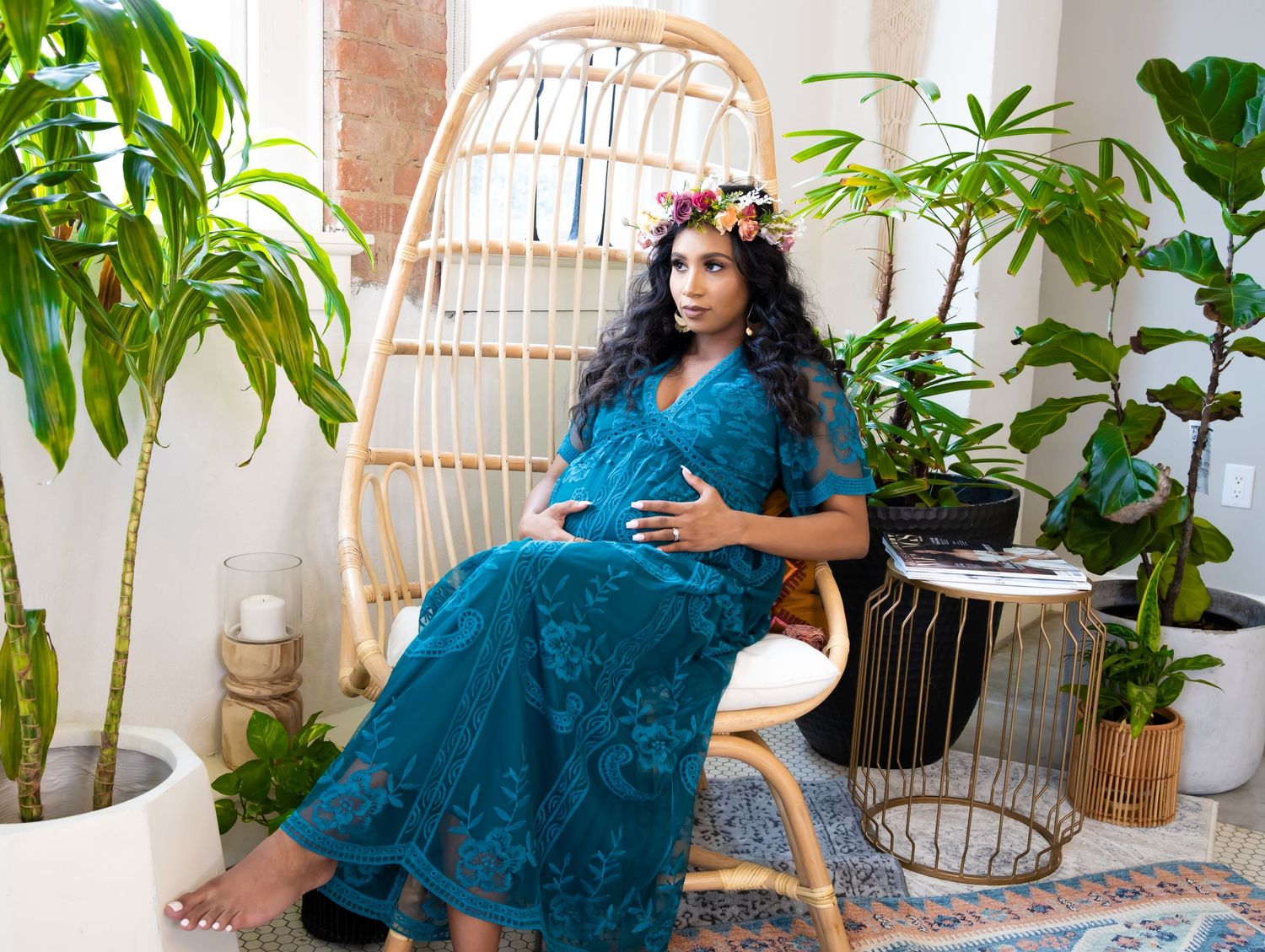 A person in a teal lace dress sits in a wicker peacock chair surrounded by tropical plants on a decorated patio.