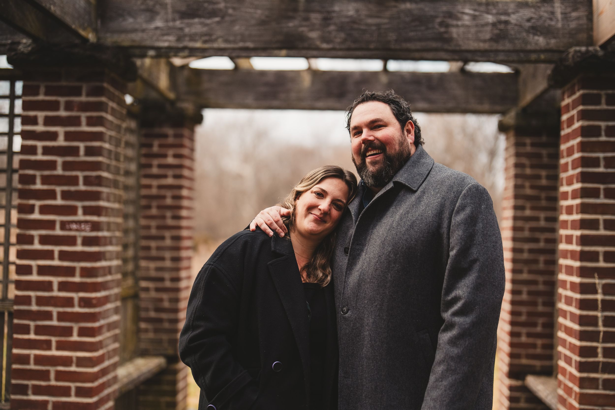 A couple in winter coats share an intimate moment under a brick archway on a misty day in Georgetown, DC after their surprise proposal.