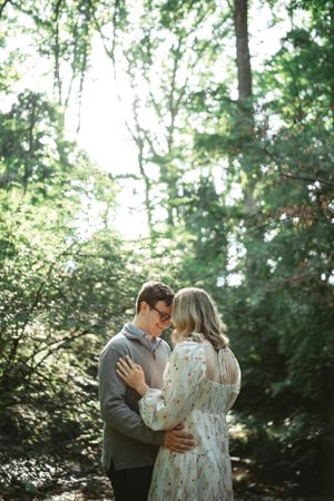 A romantic couple embraces in a sunlit forest clearing, surrounded by lush green trees and dappled sunlight in Rockville, MD.