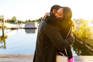 A couple embraces on a dock at sunset with boats and water in the background at the Wharf in Washington, DC after their surprise proposal.