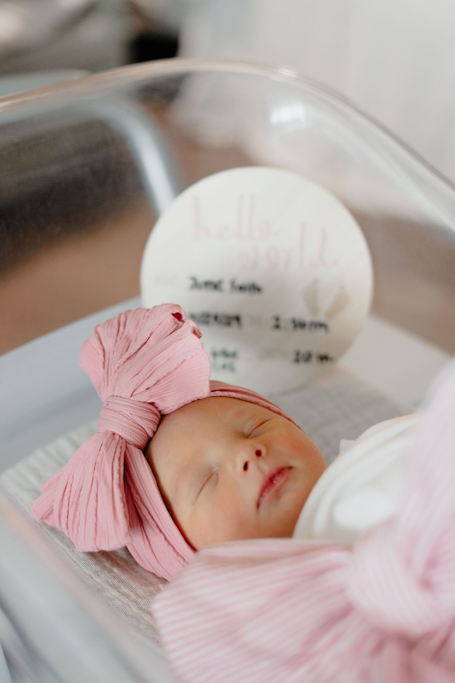 Newborn baby sleeping peacefully in hospital bassinet wearing pink bow headband.