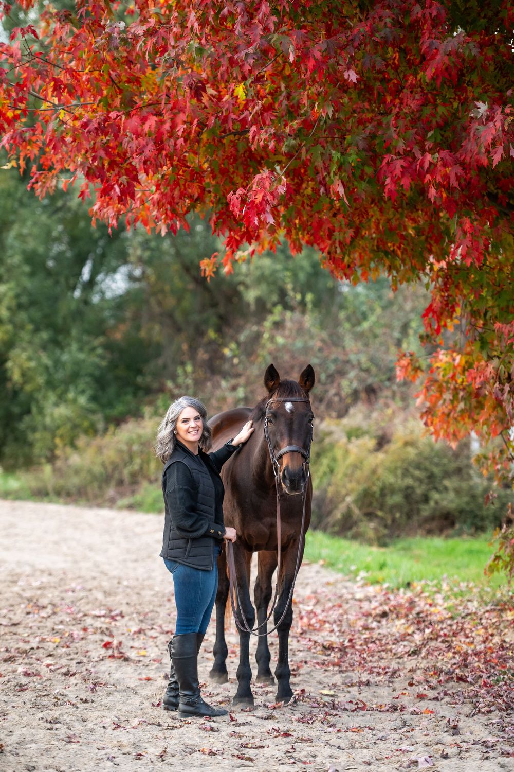 Woman with gray hair stands beside dark brown horse under vibrant red autumn foliage.