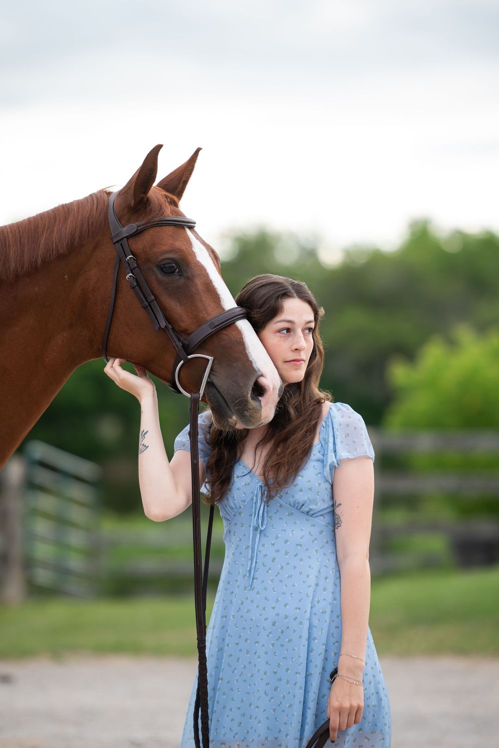 Young woman in blue dress standing beside a chestnut horse with bridle outdoors.