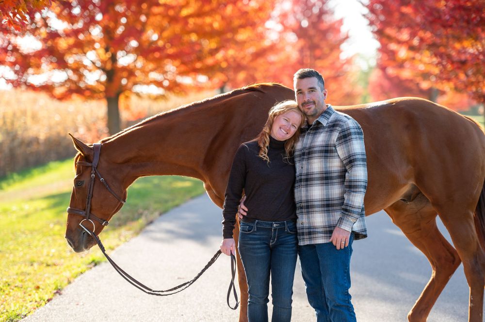 Beautiful bay horse poses with couple on autumn path with vibrant red foliage.