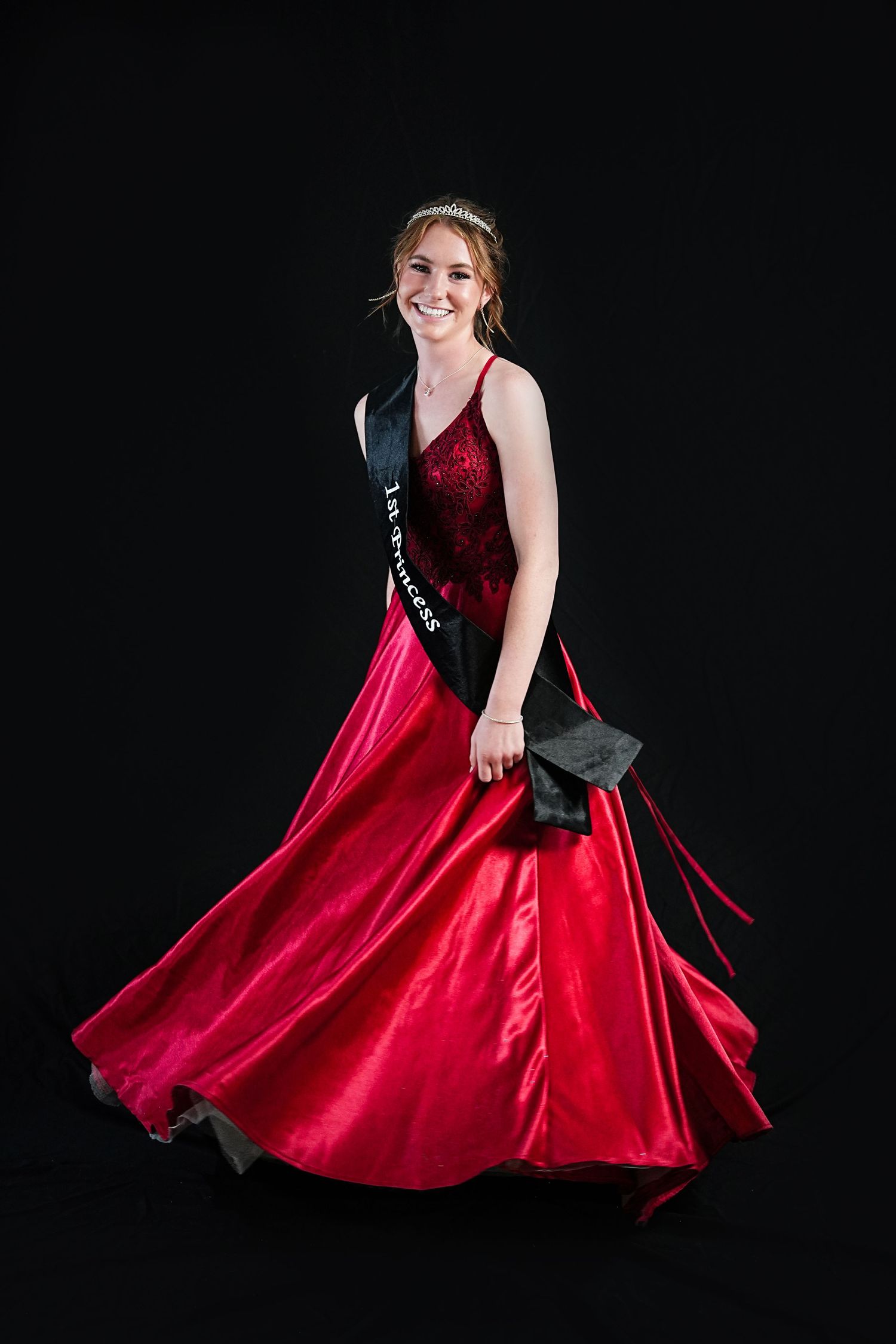 Young woman in flowing red formal gown twirling against black background.