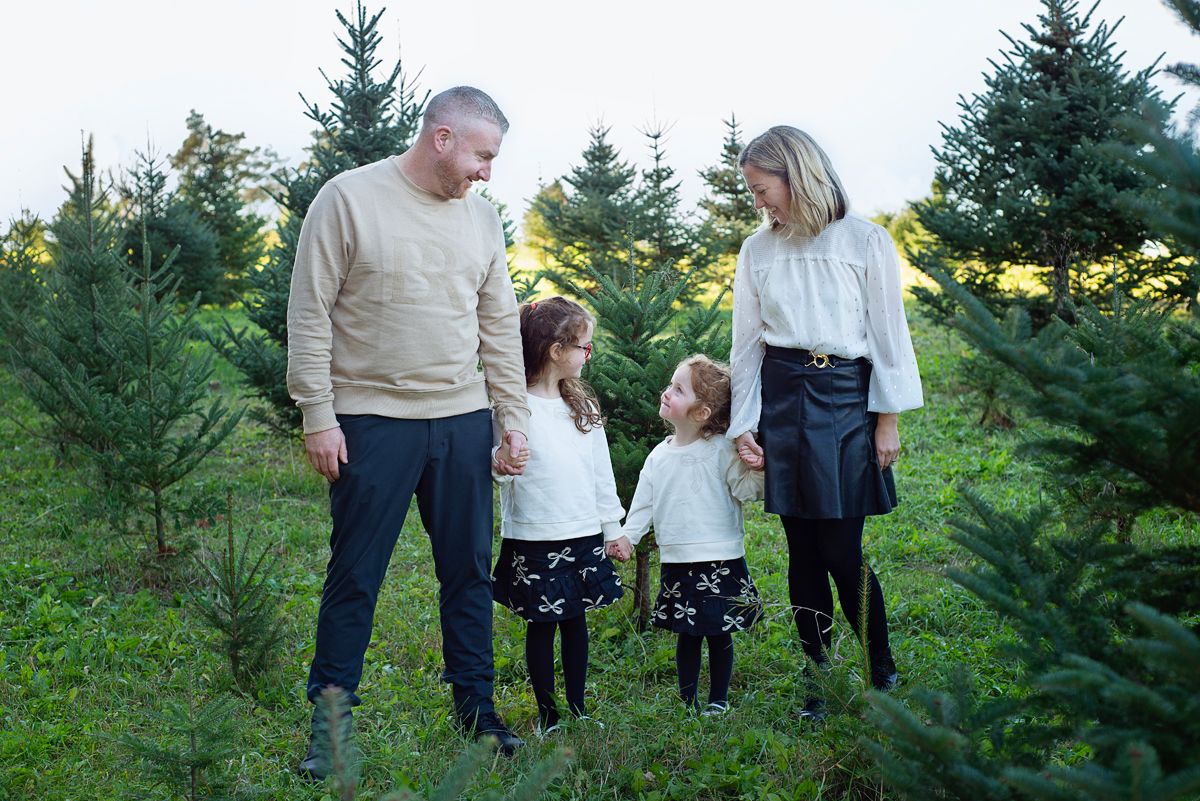 pull back of family of 4 with parents and 2 school age girls look up at each other smiling in a Christmas Tree Field