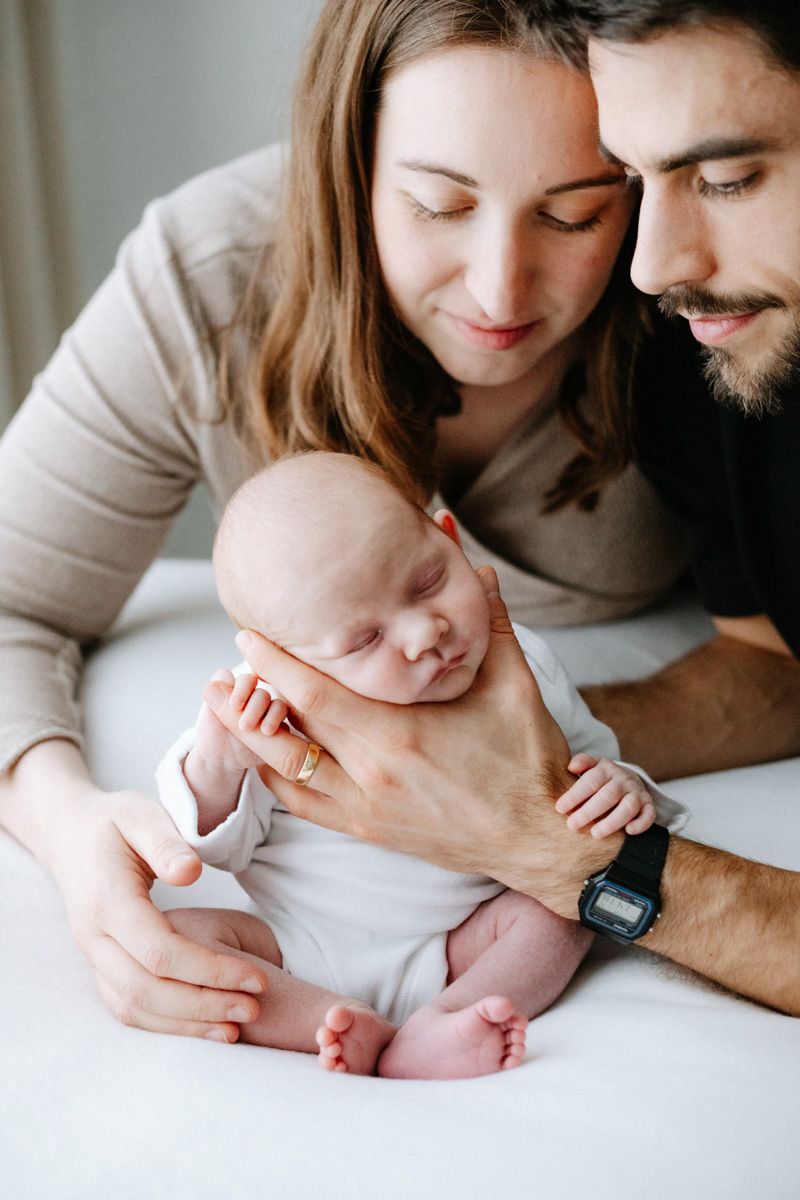 Young parents lovingly cradle their sleeping newborn baby in white outfit during an intimate family portrait.