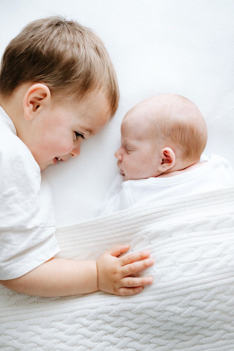 A toddler boy in white shirt smiles at sleeping newborn baby sibling wrapped in white knit blanket.