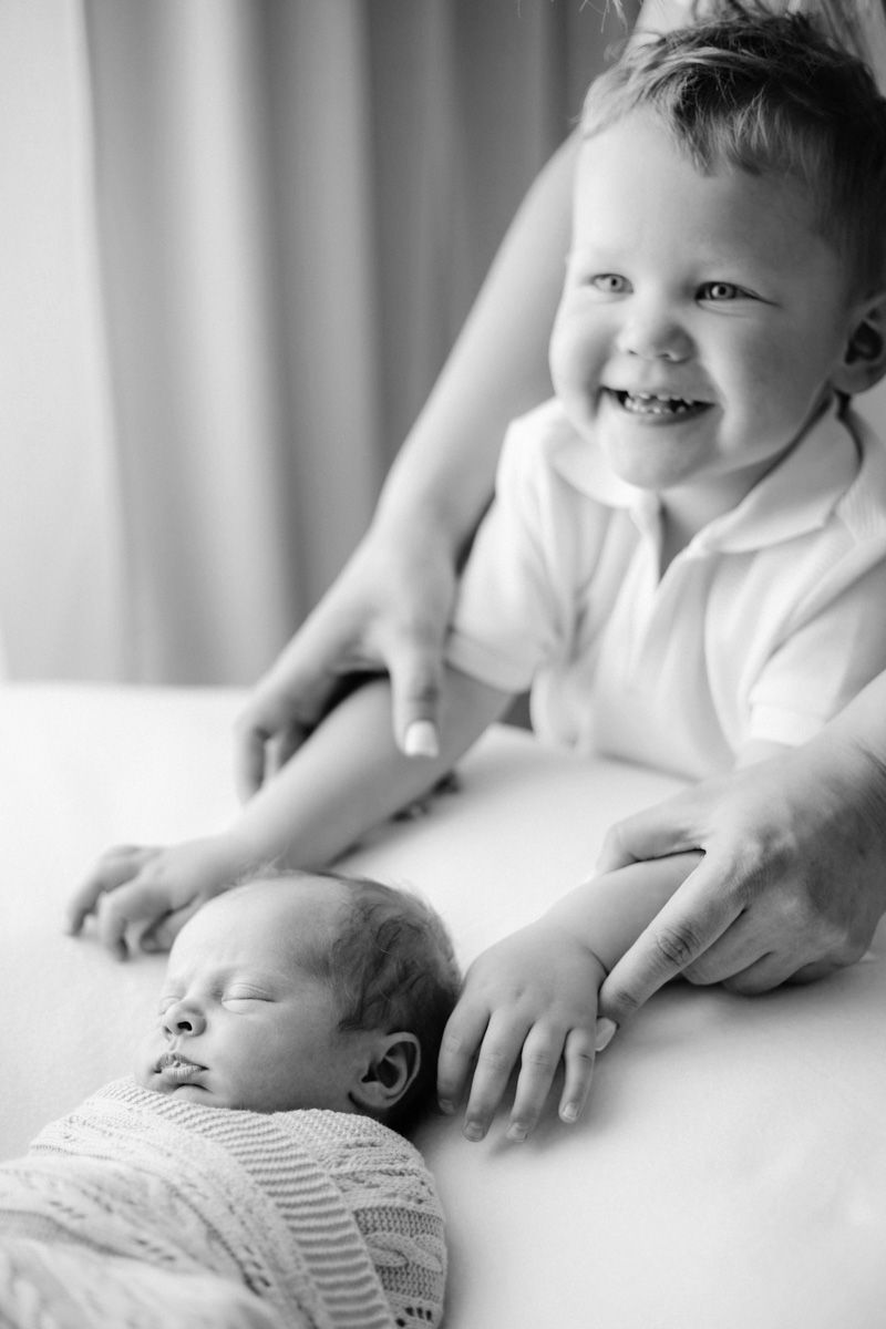Black and white photo of a smiling toddler boy gently touching his sleeping newborn sibling on a white surface.
