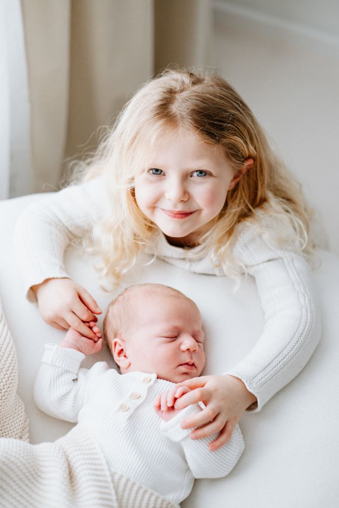 Two children in white sweaters snuggling together, one older with blonde hair and one sleeping newborn.