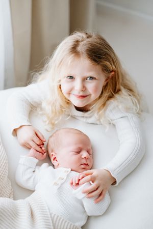 Two children in white sweaters snuggling together, one older with blonde hair and one sleeping newborn.