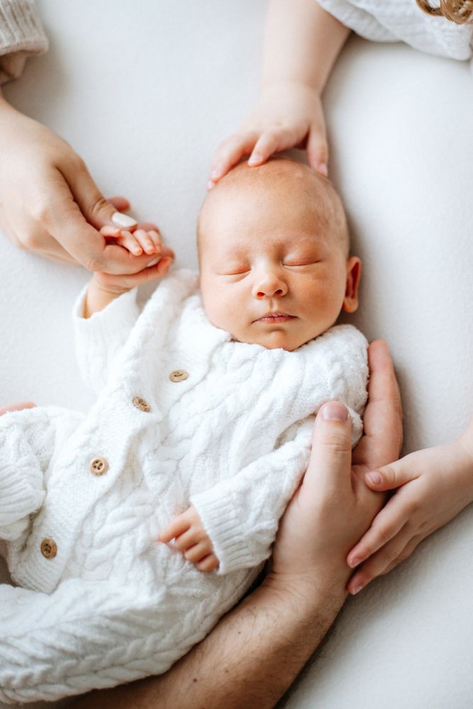 A sleeping newborn in a white knit sweater is gently held by caring hands against a light background.