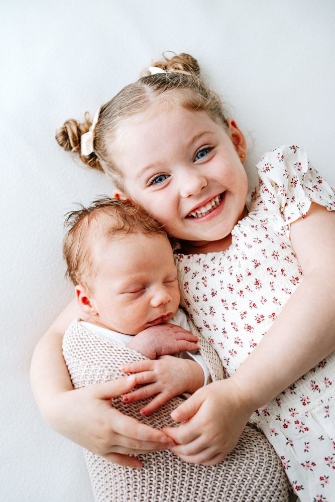 Smiling young child in floral dress gently holding a sleeping newborn baby wrapped in knitted blanket on white background.
