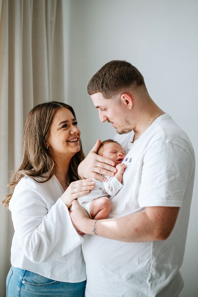 Young couple in white shirts share an intimate moment with their newborn baby against a light curtain background.