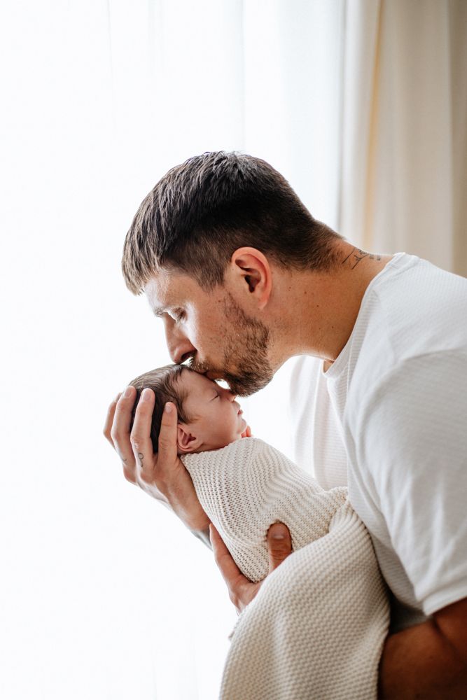 A tender moment as a parent kisses their newborn baby who is wrapped in a white knit blanket near a window.