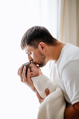A tender moment as a parent kisses their newborn baby who is wrapped in a white knit blanket near a window.