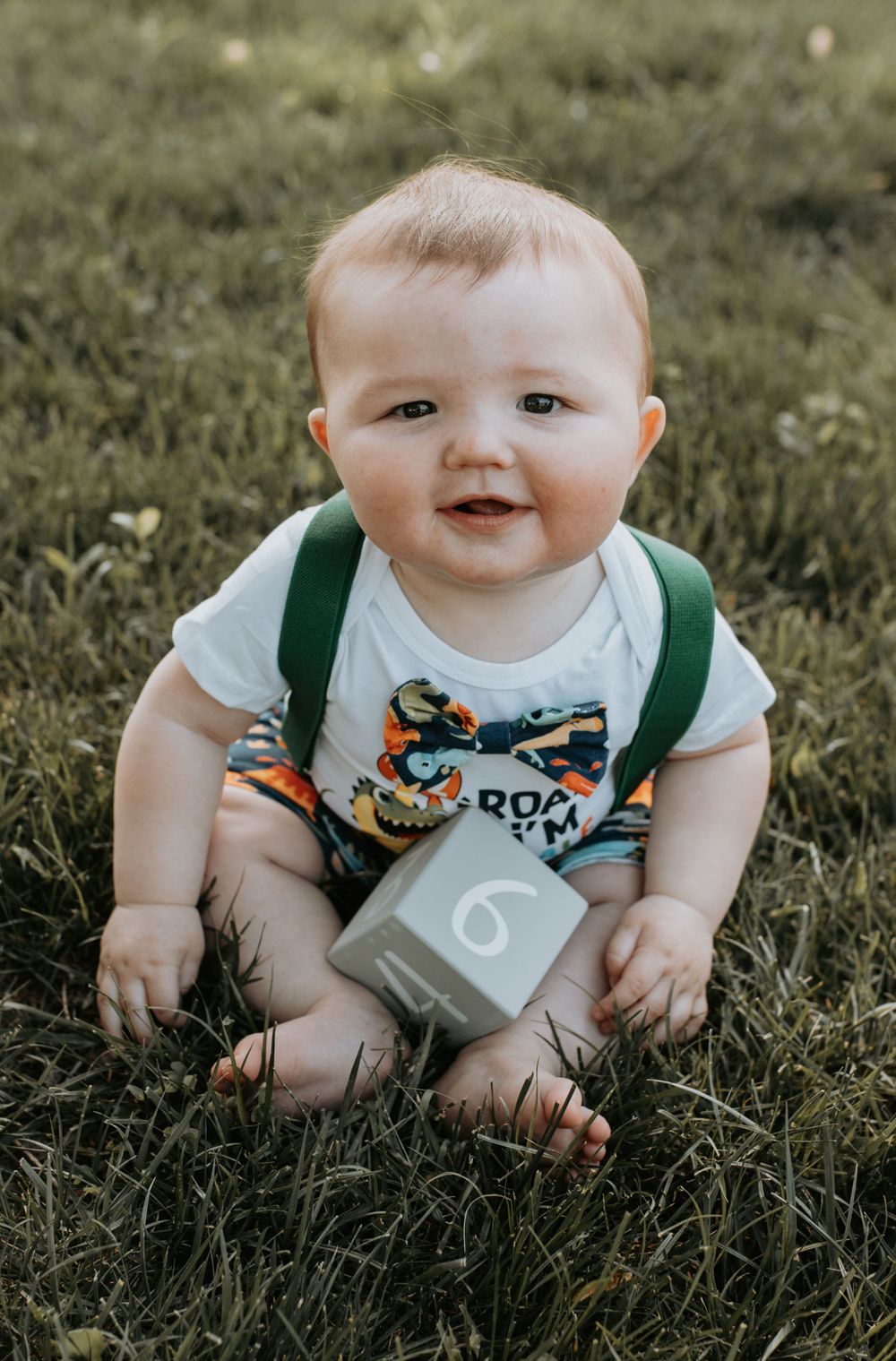 Baby wearing green overalls and white shirt sits on grass with wooden block.