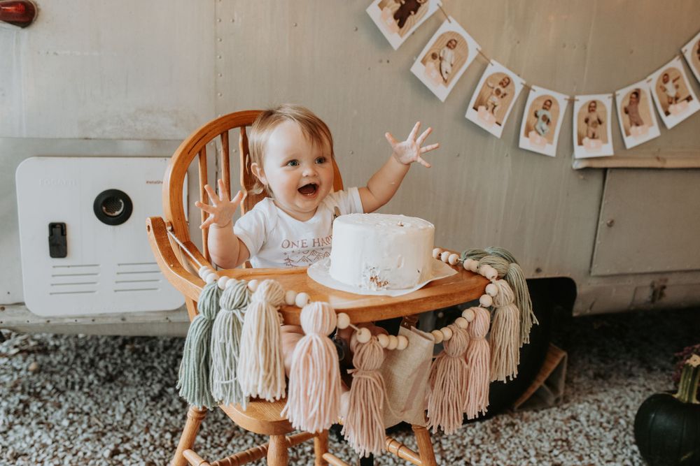 Wooden high chair decorated with tassels and a white birthday cake against light wall.