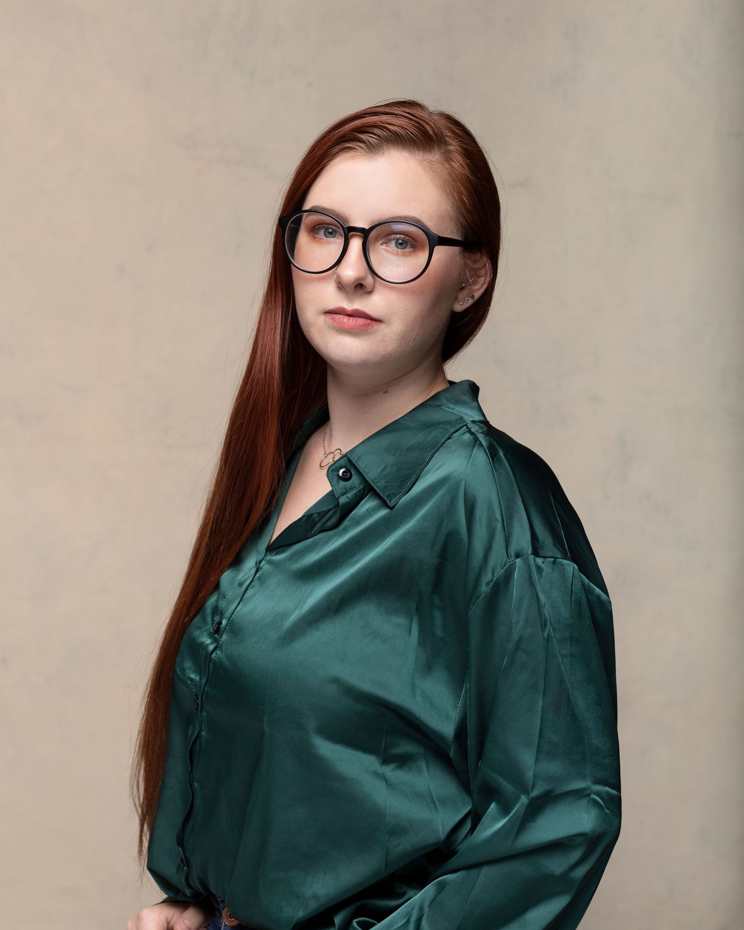 Young woman with long red hair and round glasses wearing a green satin blouse, posing against a neutral background.