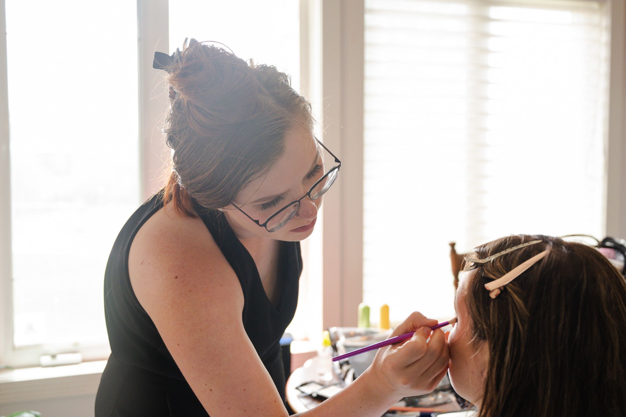 Professional makeup artist applying bridal makeup to a client near a bright window.
