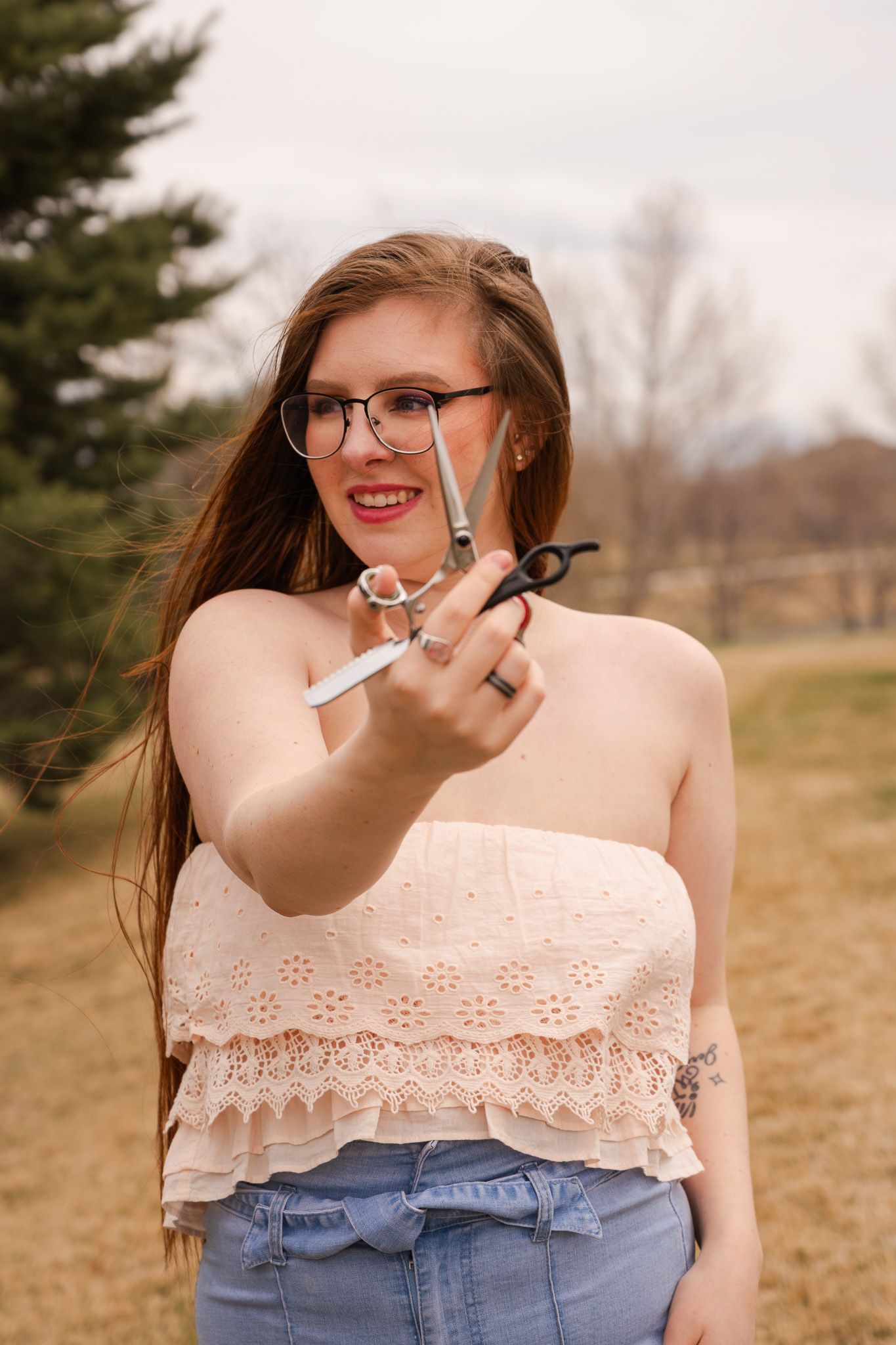 Young woman with glasses and red lips holds scissors toward camera outdoors wearing a strapless top and jeans.