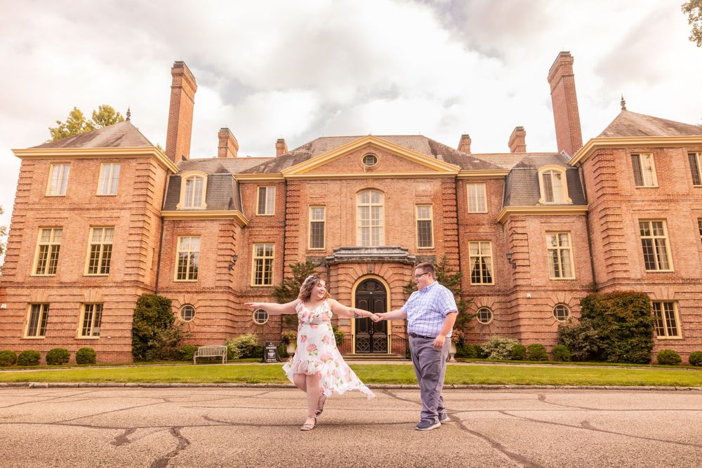 A couple walks hand in hand in front of a grand pink brick mansion with classical French architecture.