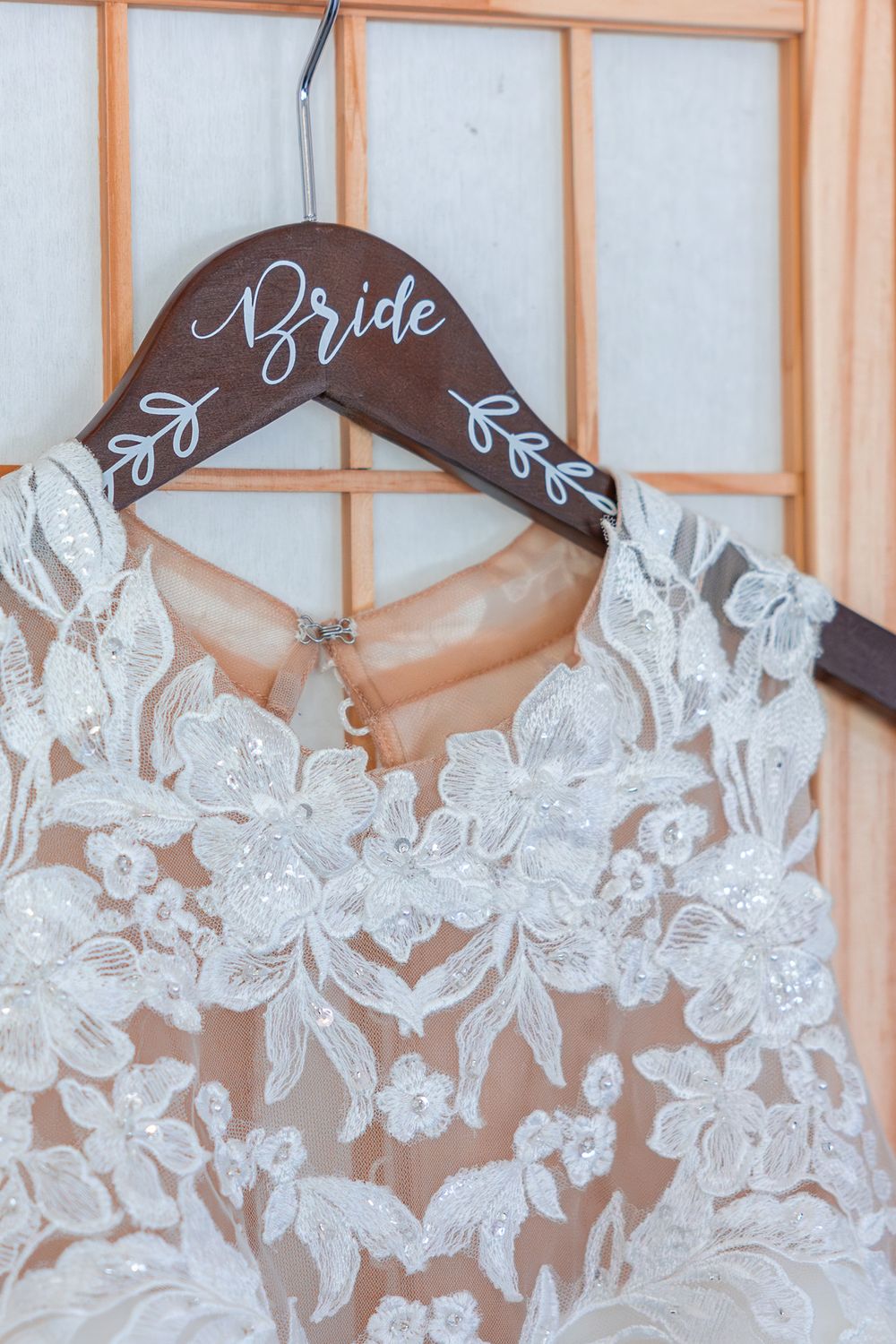 Wedding dress with delicate lace floral details hangs on a wooden hanger labeled 'Bride'.