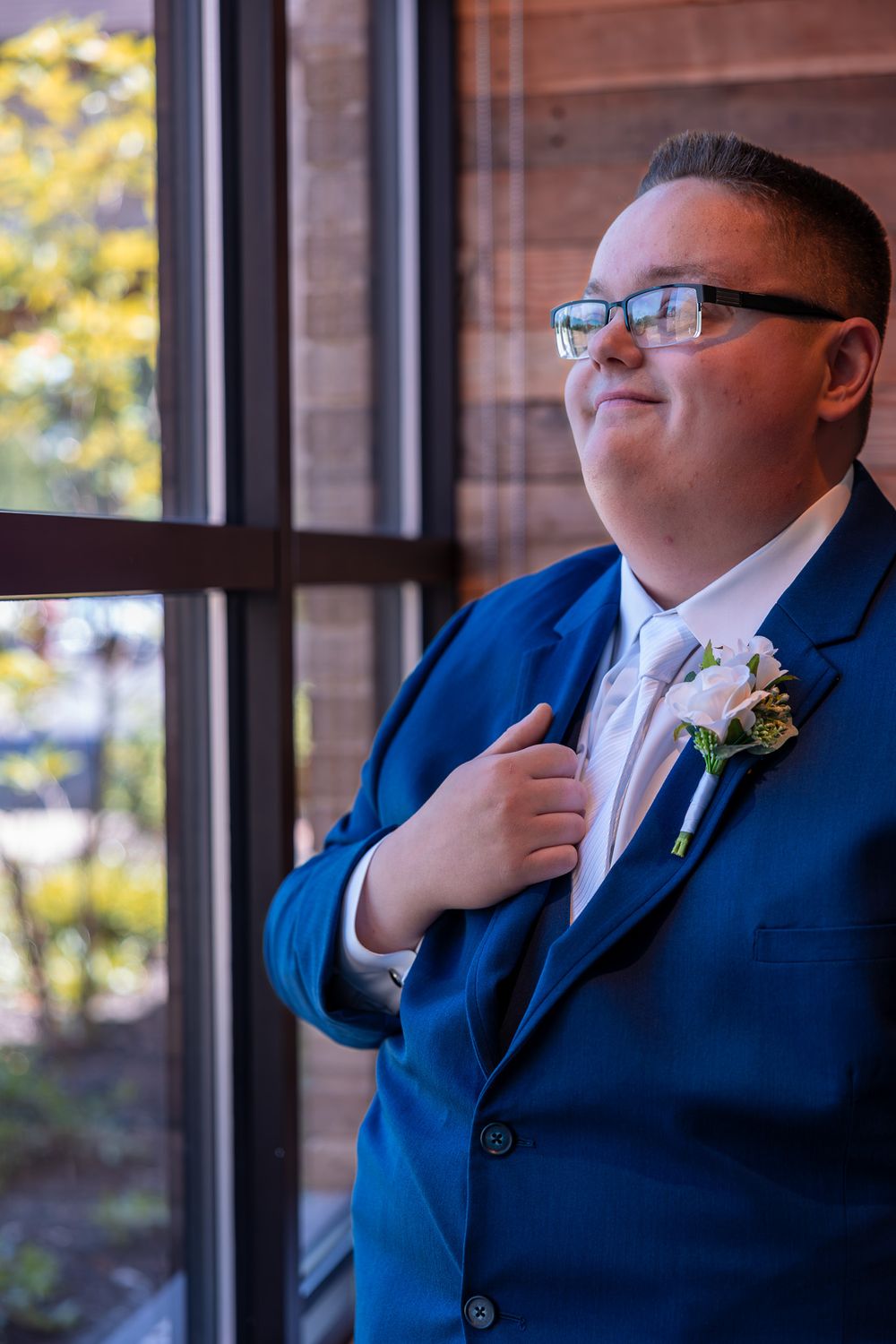 A groom in a bright blue suit and glasses stands thoughtfully by a window during a wedding celebration.