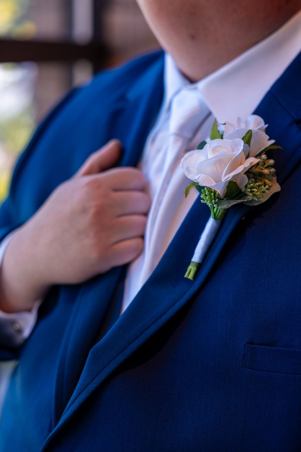 A delicate pink rose boutonniere adorns a navy blue suit jacket with white shirt and light blue tie.