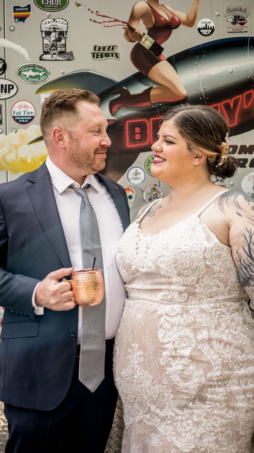 A couple in wedding attire share a joyful moment in front of a decorated wall as the groom holds a copper mug.