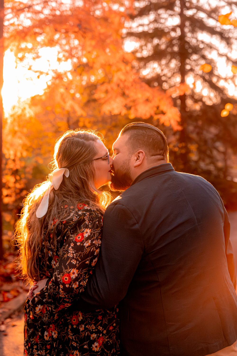 A couple shares a romantic kiss at sunset with vibrant orange autumn foliage glowing in the background.