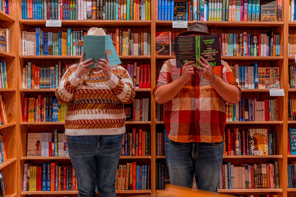 Two people stand in a library holding open books in front of their faces against a backdrop of colorful bookshelves.