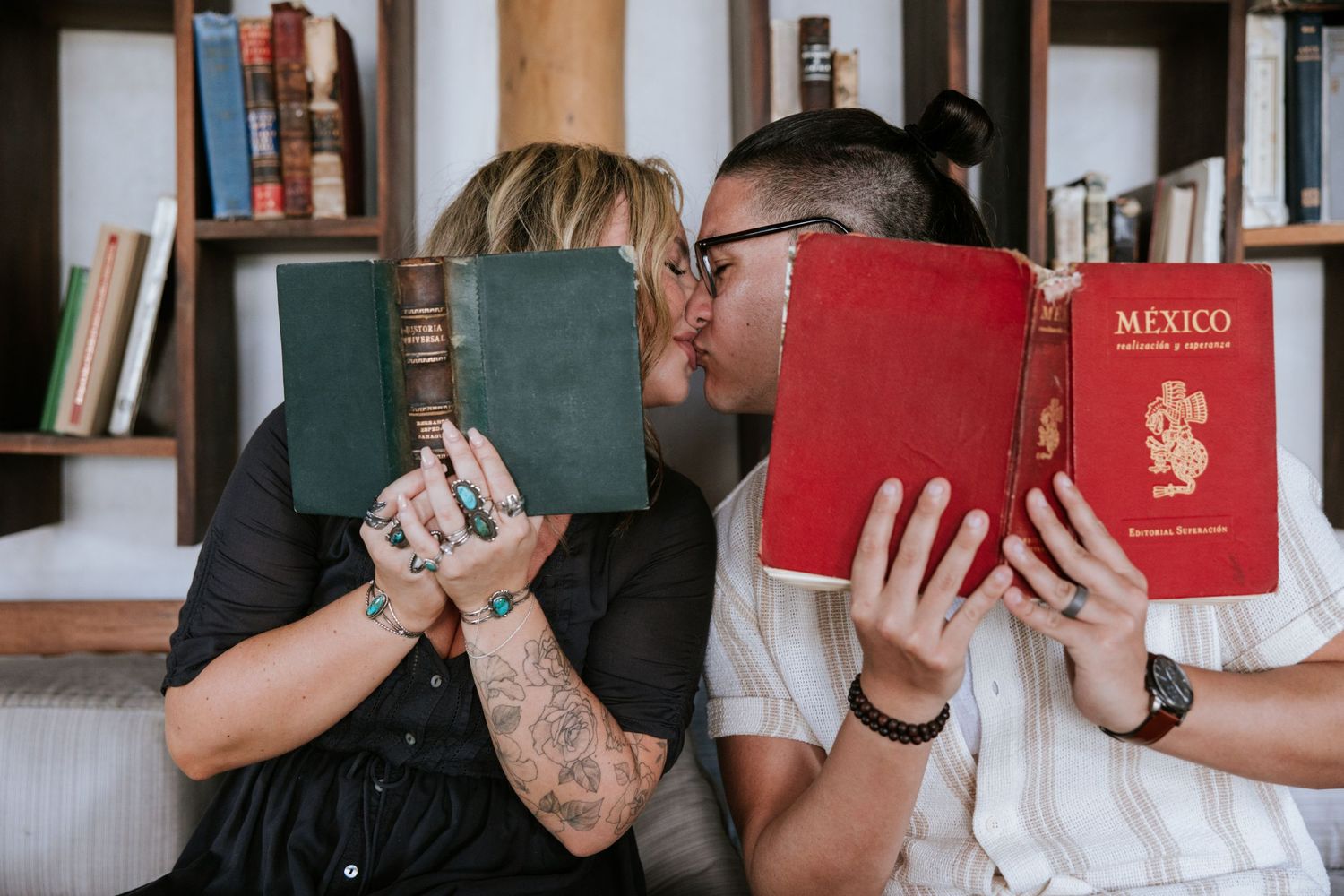 Engagement portrait of a couple in a cozy library, playfully hiding behind books for a secret kiss