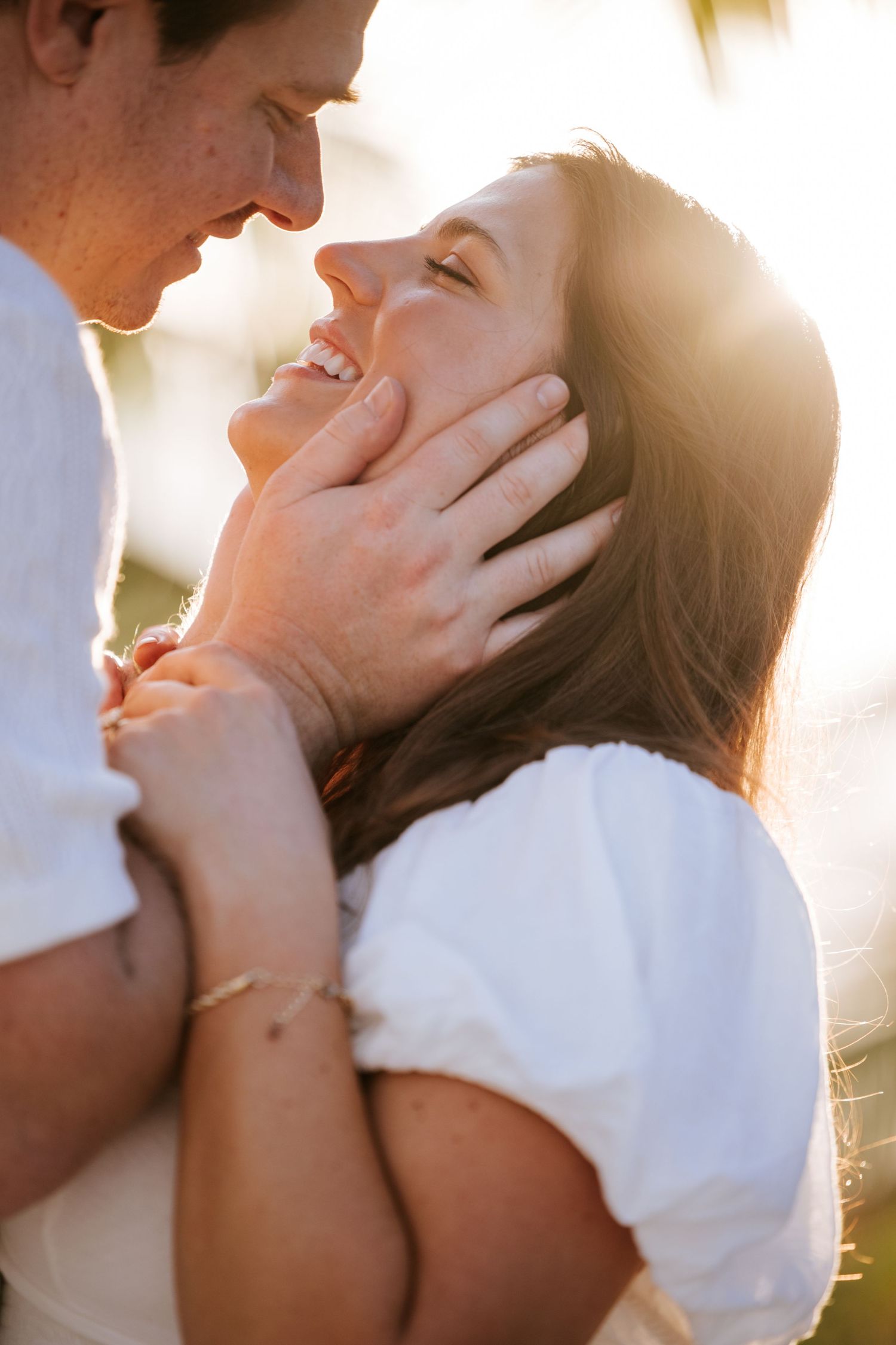 Close-up of a couple at sunset, as he gently holds her face, bringing her closer for a kiss, with golden sunlight glowing between them