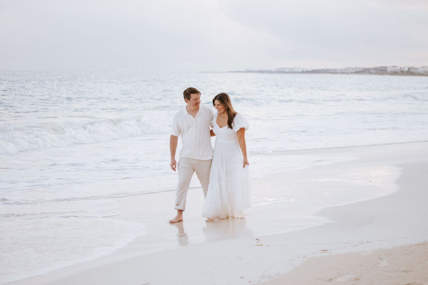 Couple walking hand in hand along the shore in Costa Mujeres, splashing water and laughing as the waves touch their feet at sunset.
