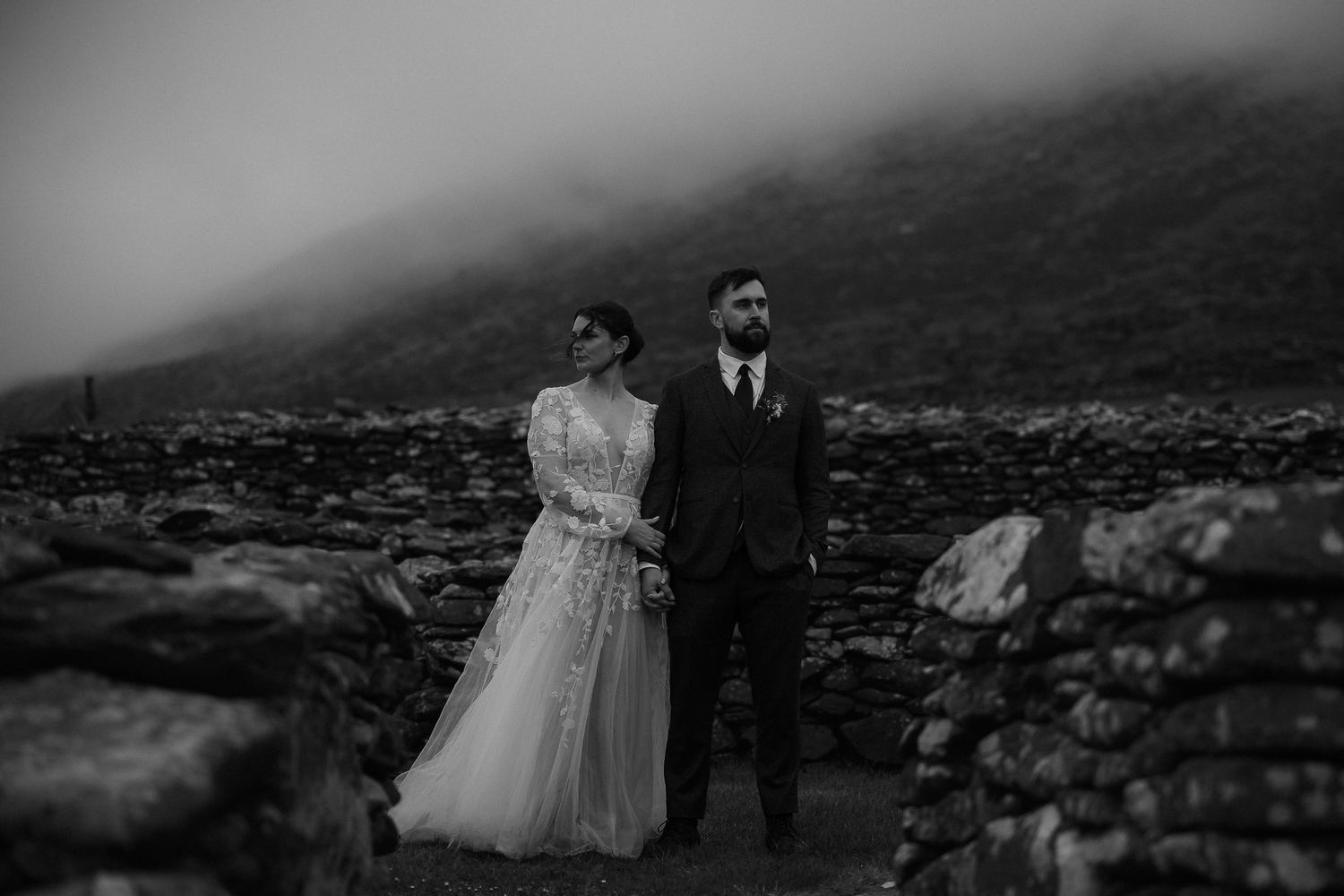 A romantic black and white wedding portrait taken against a moody mountainous backdrop with stone walls.