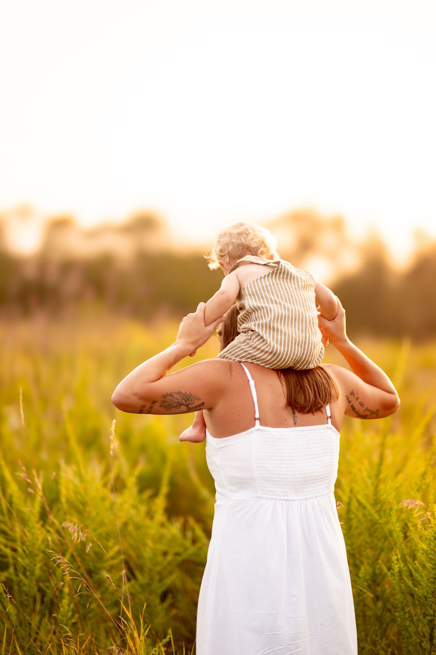 A person wearing a white sundress carries a small child on their shoulders in a sunlit field at golden hour.