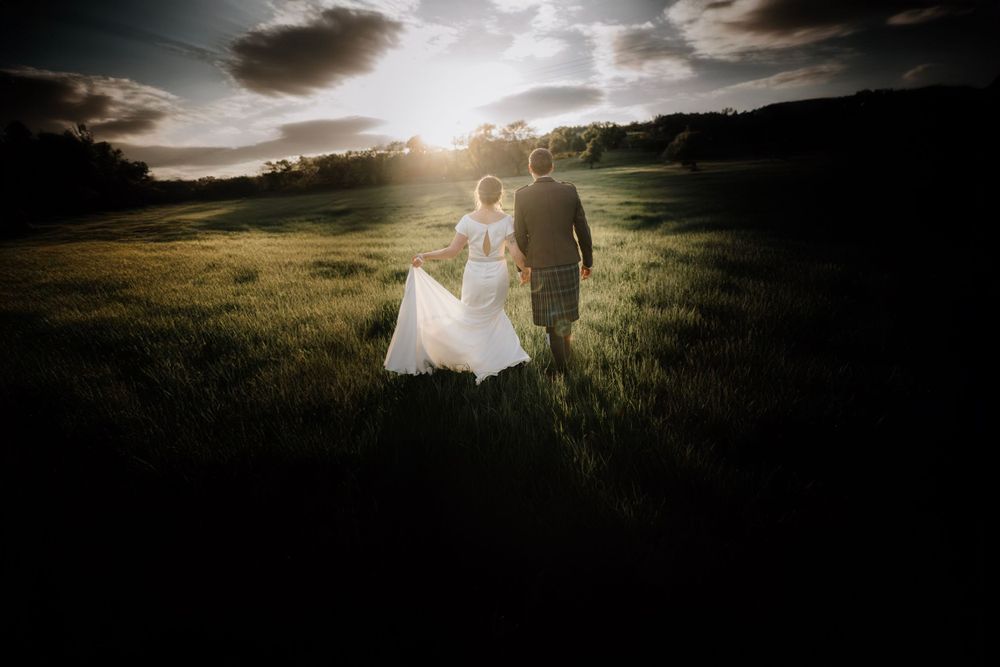 Silhouette of newlyweds walking in a sunset-lit meadow with dramatic sky.