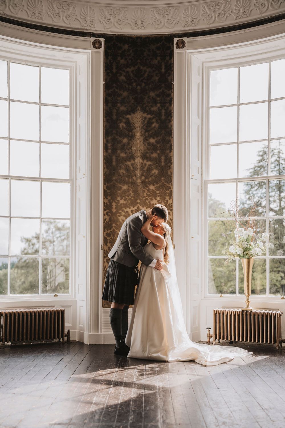 A romantic wedding moment captured in an elegant bay window setting with natural sunlight streaming in.