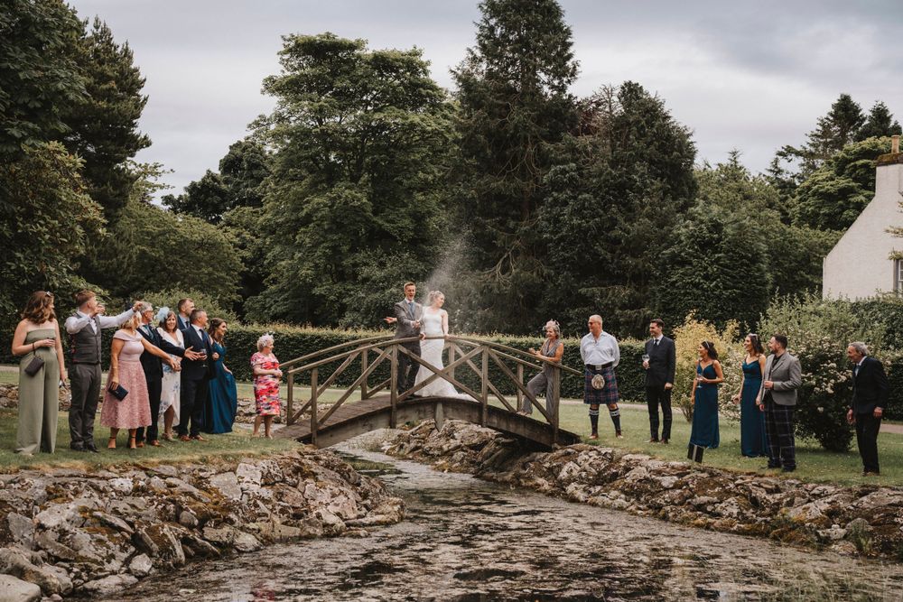 Wedding party gathered on rustic wooden bridge for outdoor photo.