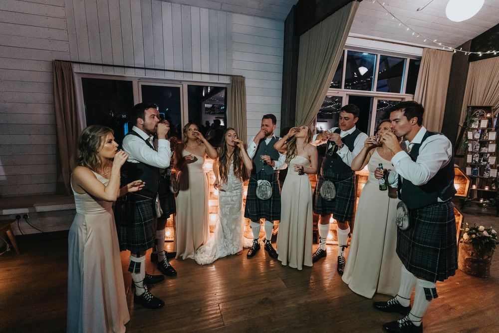 Wedding guests dance in traditional Scottish attire during an evening reception with string lights.