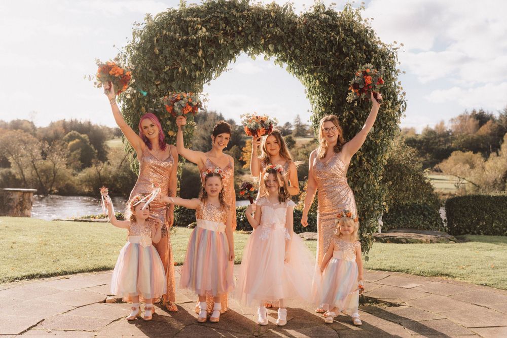 Bridesmaids in pink dresses celebrate joyfully under a floral arch in an outdoor garden setting.