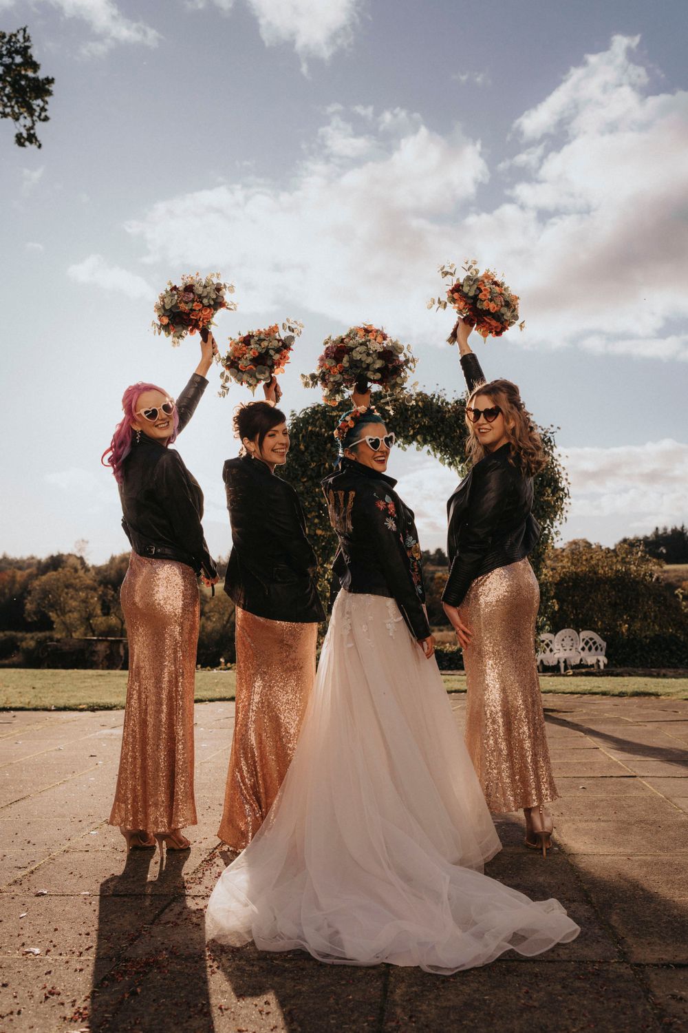 Bridal party in sparkly dresses holding bouquets against cloudy sky.