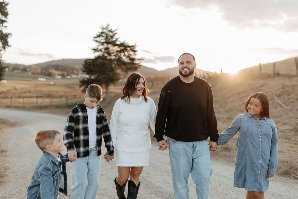 Family holds hands while walking on rural path during golden sunset.
