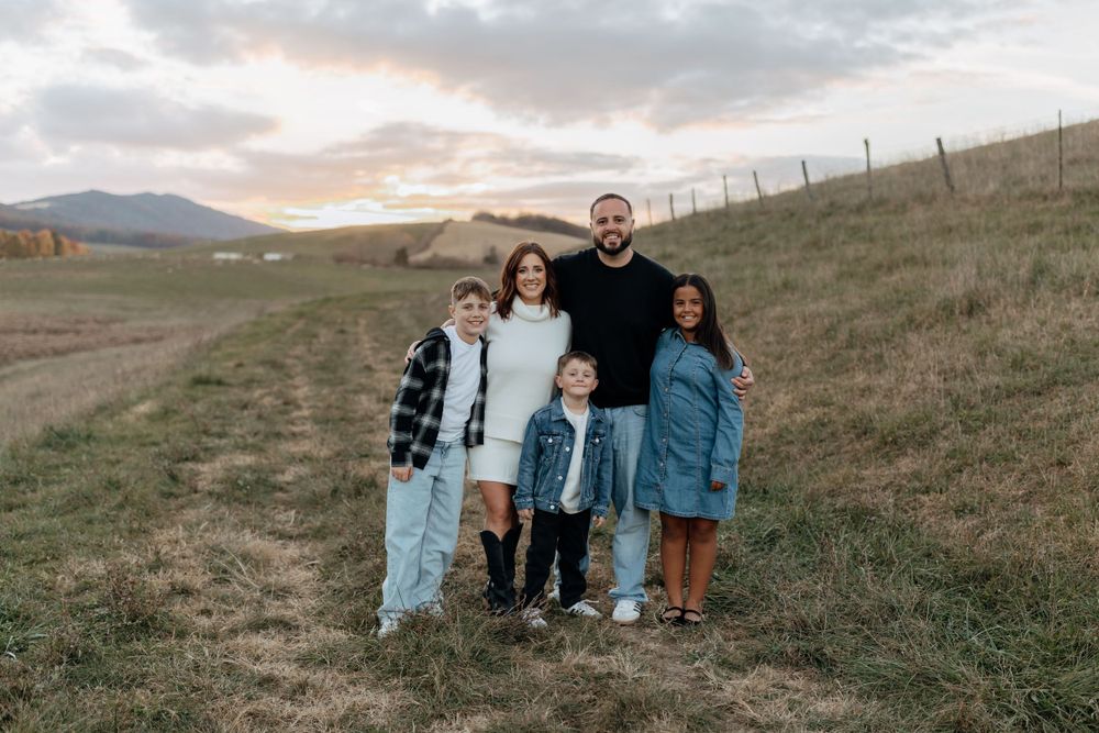 Family in coordinated casual outfits poses together in mountain field at sunset.