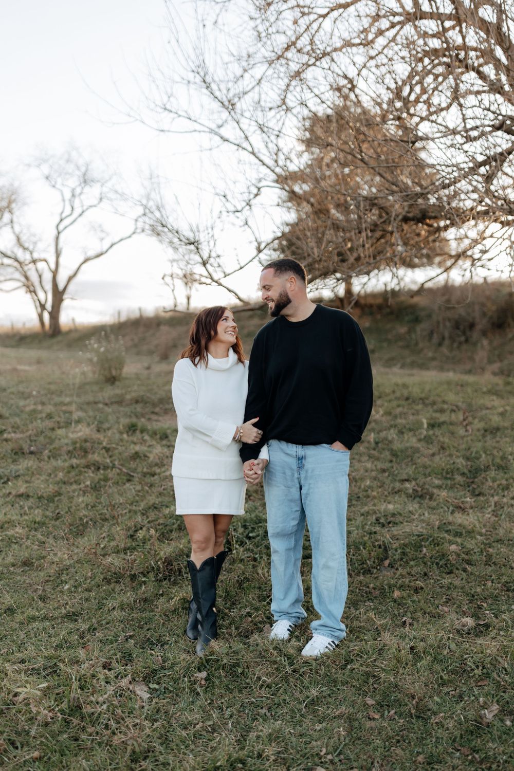 Couple in casual wear standing together on grassy hillside at sunset.