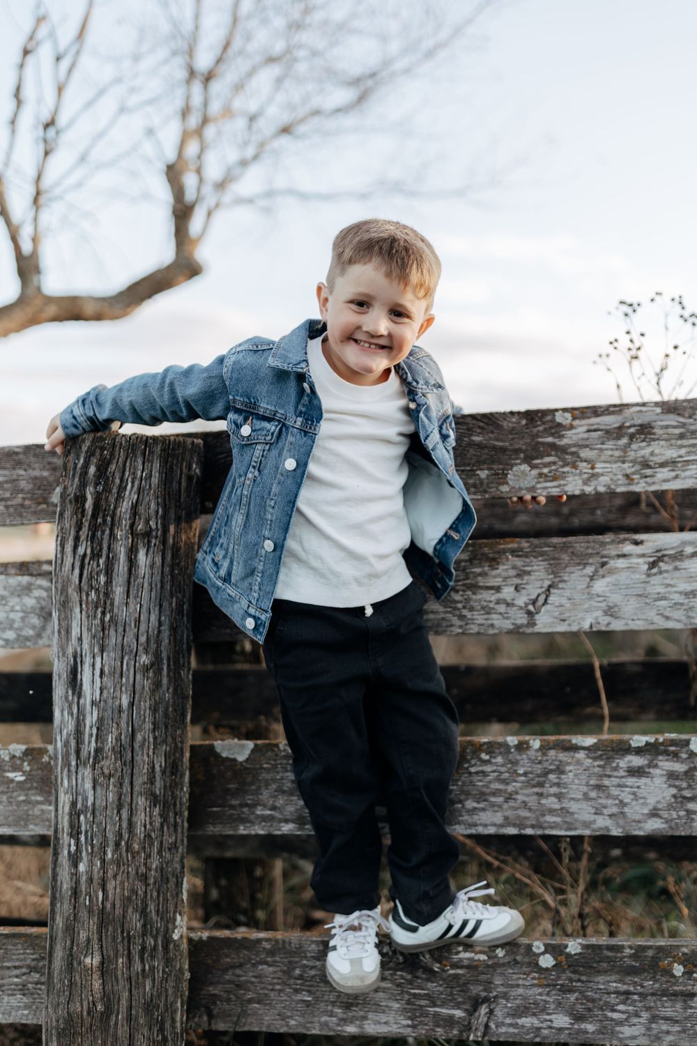 Child in denim jacket leans casually against rustic wooden fence post on a bright day.