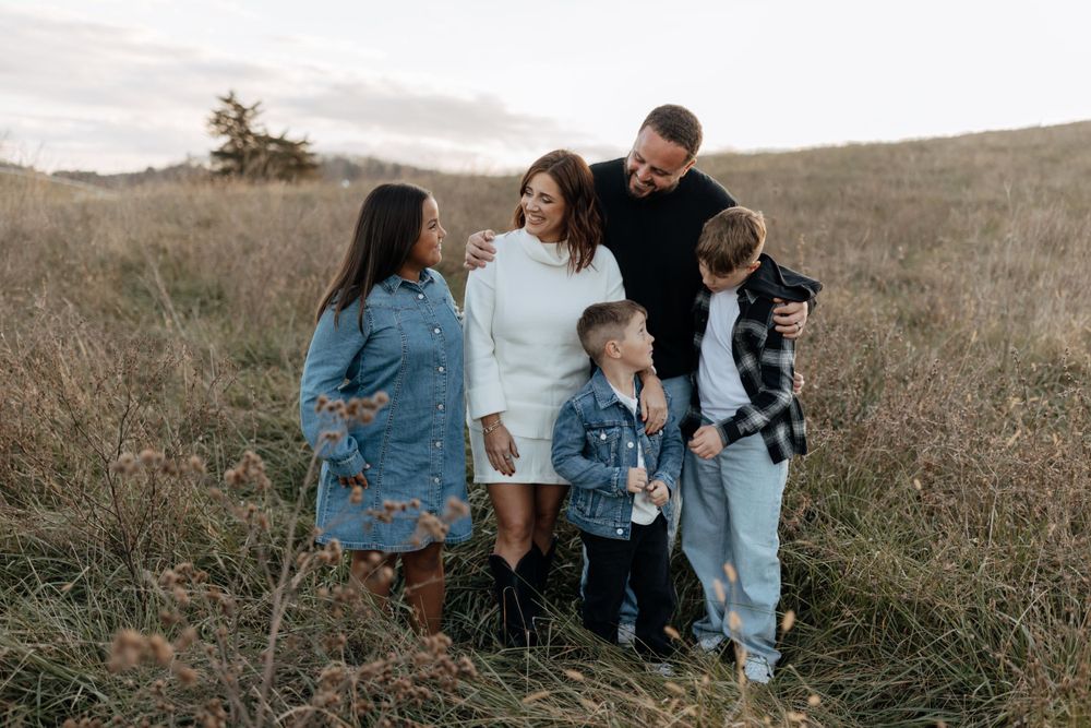 Family wearing coordinated blue and white outfits stands together in golden field at sunset.