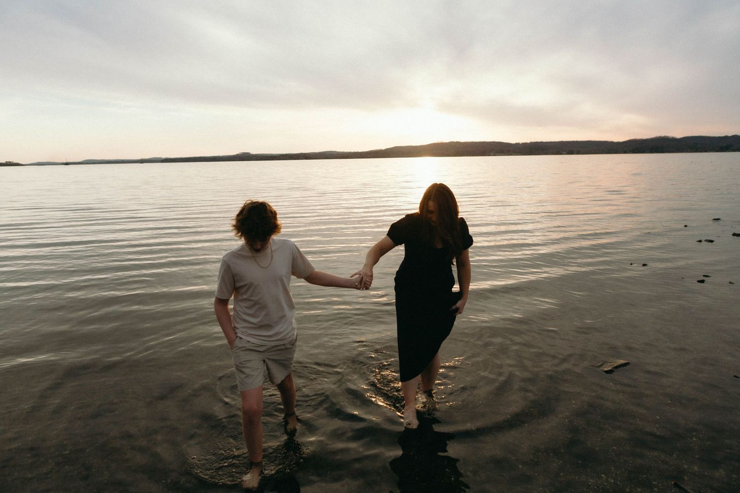 Two people holding hands while walking along a beach at sunset with calm waters and distant shoreline in view.